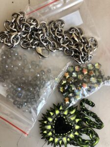 photo of stainless steel chain next to two baggies of beads, one small moon quartz and the other small starts in greens and oranges. bottom right corner is a plastic shape of a spiky heart with chains